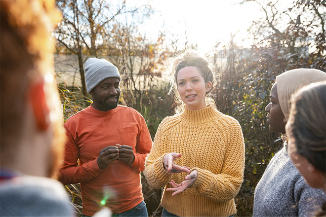 Lady talking to a group of people standing outside infront of bushes