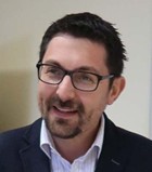 A headshot of Prof. Yannis Tsalavoutas, dressed in a dark suit and white shirt, against a blurred background. He is in mid-conversation.